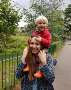 woman with long brown hair and child on her shoulders, the child has orange welly boots and a red jacket on, and has blonde hair.