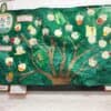 3D image of a handmade tree noticeboard from a school with a green background and brown tree and branches covered in apple shaped cards.