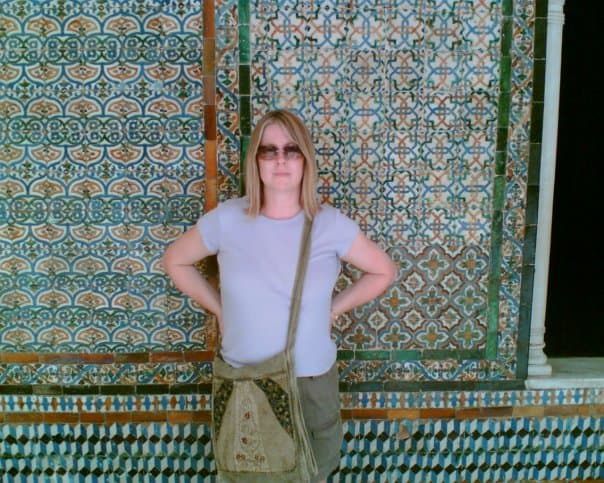Woman with mid brown hair in front of tiled wall wearing sunglasses and a purple t-shirt