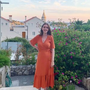 Woman with brown hair and long orange dress on standing at the top of steps with a city view behind her