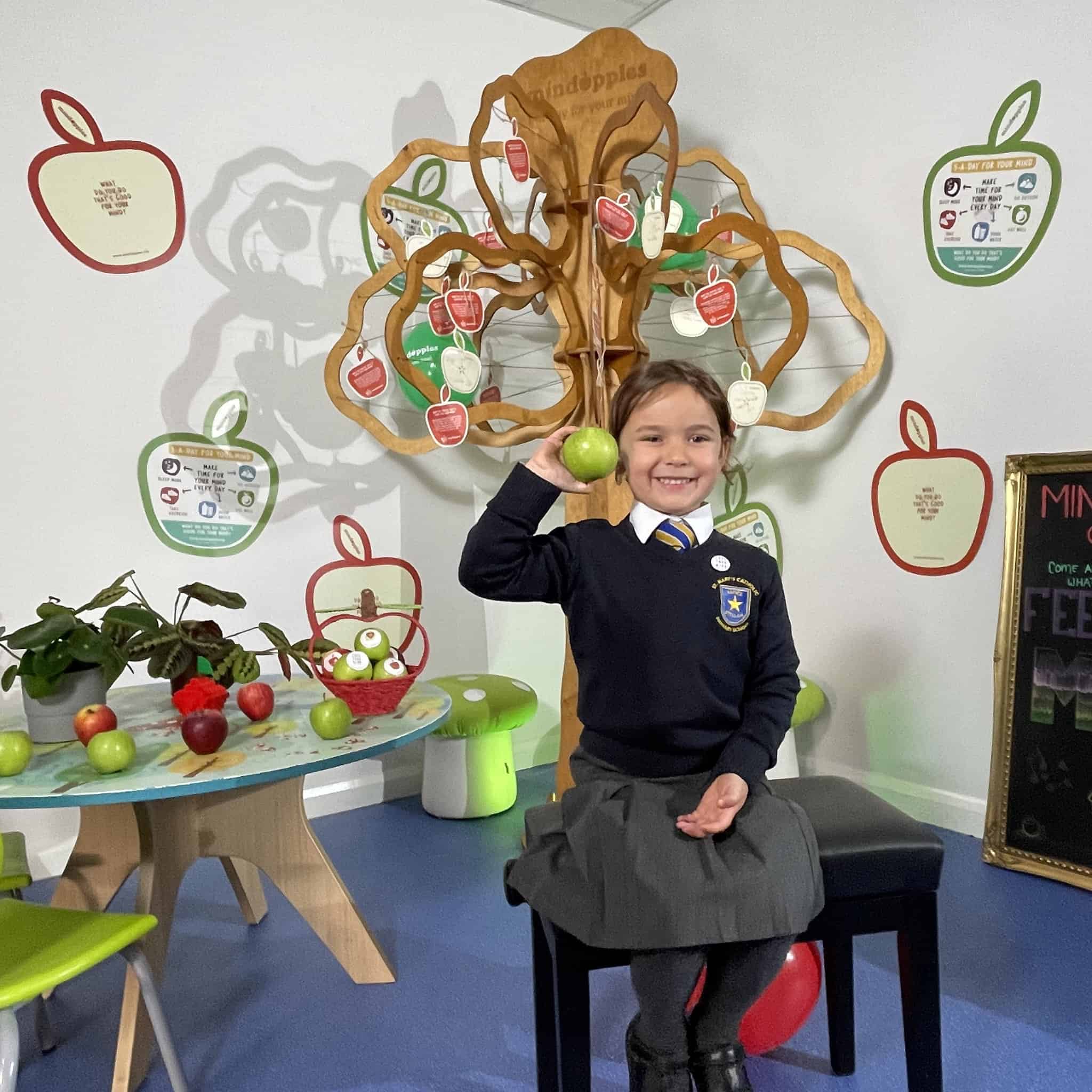Schoolgirl smiling holding up a green apple sat in front of a wooden tree with hanging apple shaped applecards. Mindapples promote positive mental health from an early age.