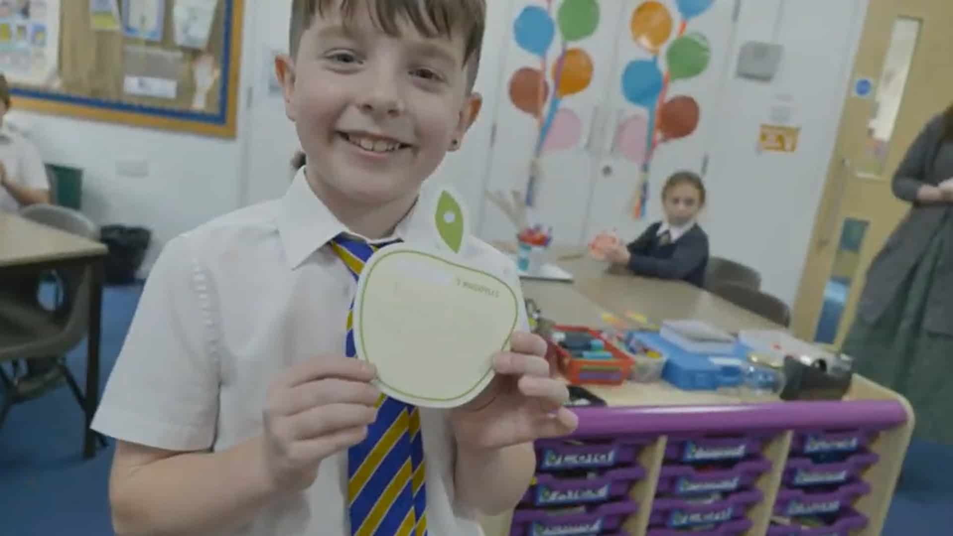 Smiling boy in classroom holding up his apple shaped card where he has written 5 things he does every day to look after his mind. Mindapples promote positive mental health from an early age.