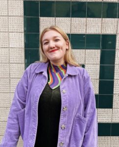 Blonde woman wearing a purple coat, standing in front of green and white tiled wall.