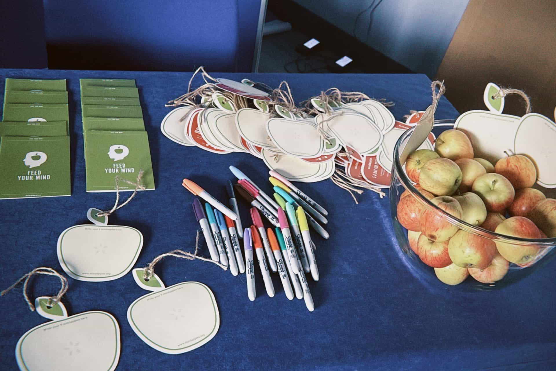 Planned table with Mindapples signs, colourful markers, and a bowl of apples promoting mental wellbeing and mindfulness activities.